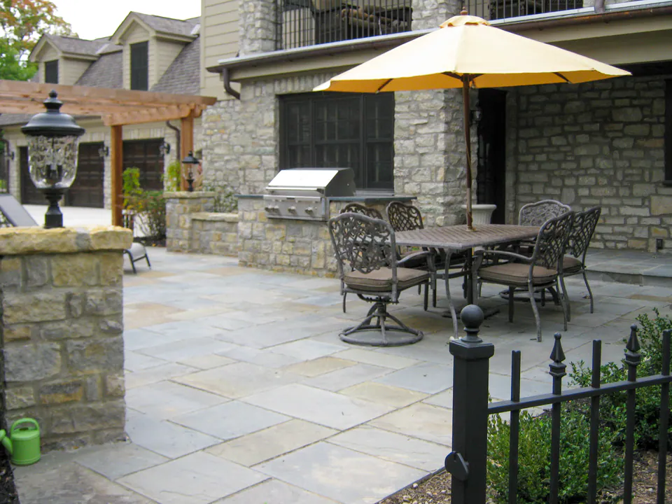 Bluestone patio with built-in grill, dining table, umbrella, stone wall, and garden planting